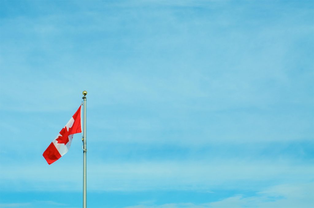 Flag pole with raised Canadian flag set in from of a blue sky backdrop.